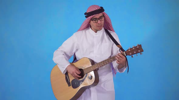 Arab Man Clumsily Plays Acoustic Guitar on Blue Background alt