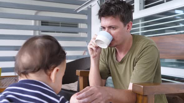 The Father is Sitting on a Wooden Chair and Drinking Coffee From a Mug His Son is Playing in Front alt