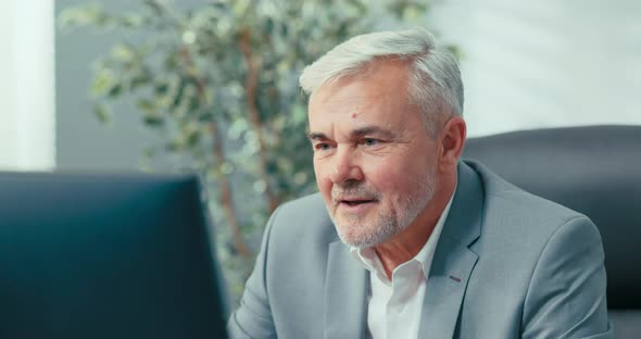 Elegant Man in Suit with Gray Hair Conducts Remote Conference From Office Sitting at Desk in Front alt