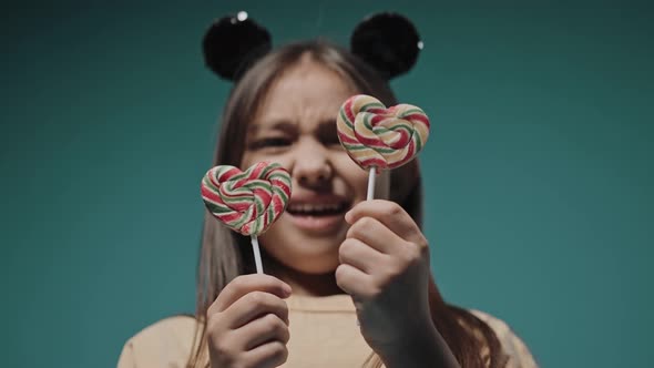 Little Girl is Choosing Between Two Heart Candies on Stick on a Blue Background