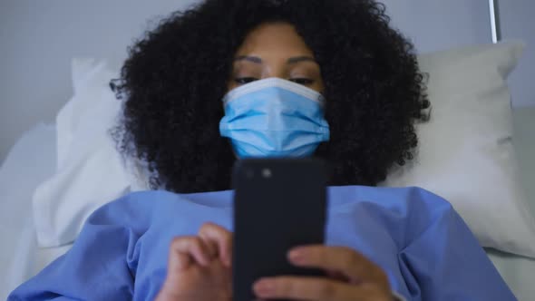 African american female patient wearing face mask lying on hospital bed using smartphone alt