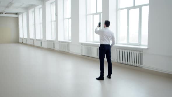 Male Realtor Stands in Middle of an Empty Office Space and Shoots It on Smartphone Back View alt
