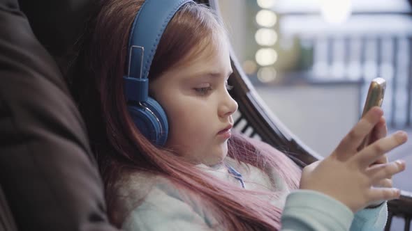 Side View Close-up of Sad Little Girl in Headphones Using Smartphone Indoors. Portrait of Upset alt