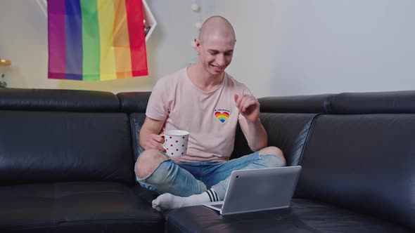 Happy Smiling Homosexual Bald Caucasian Man Sitting on a Black Couch Holding a Cup of Tea or Coffee alt