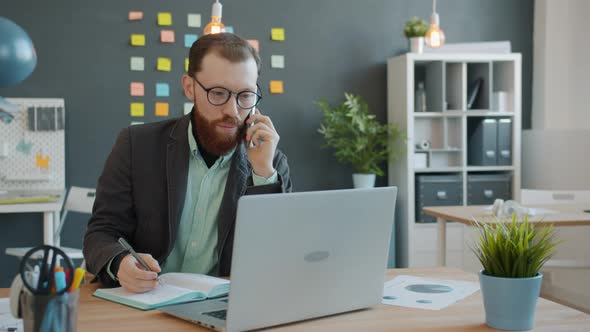 Cheerful Guy Working in Office Making Mobile Phone Call and Using Laptop Taking Notes in Notebook alt