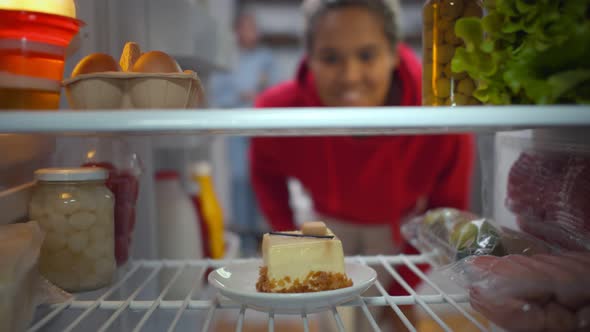 Woman on Diet Opening Refrigerator Taking Cake While Hand From Inside Fridge Holding Plate. alt