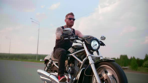 Stylish Biker with Tattoos Rides a Motorcycle on a Country Road at Sunset