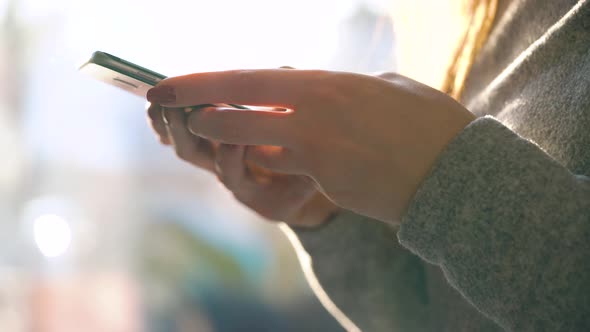 Female Hands Using Smartphone Against a Blurred Cityscape in the Setting Sun alt