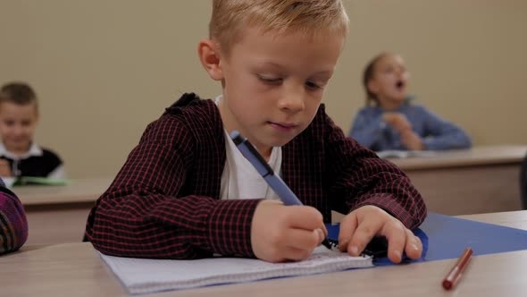 Closeup of a Little Boy Sits at a Desk in School and Writes in a Notebook alt