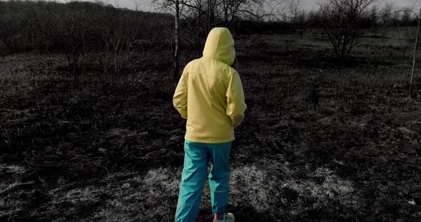 Teenage homeless girl in colorful Ukrainian clothes walks on a burnt field alt