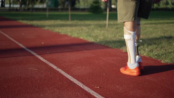 Male Amputee with Prosthetic Legs Walking on Red Sports Track in Summer Park alt