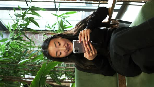 Korean Girl Sitting By The Window With Mobile Phone In Her Hands alt