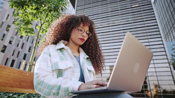 Young Female Freelancer with Afro Hairstyle is Sitting Outdoors on Street in the City in Business alt