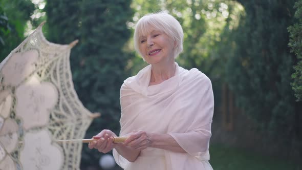 Zoom in to Happy Senior Woman in Wedding Dress Spinning Sun Umbrella Turning to Camera Smiling alt
