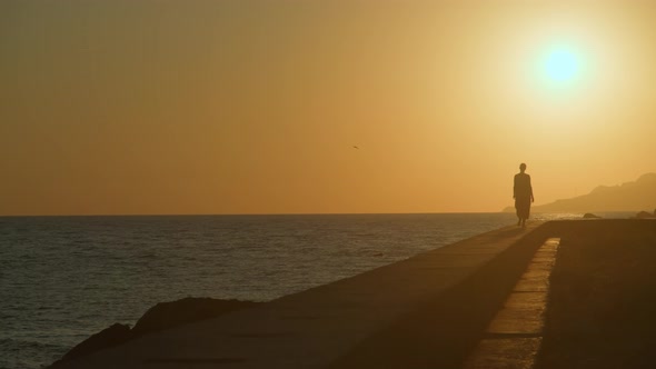 Woman in a Red Dress and a Straw Hat Walks Along the Edge Front of a Cliff By the Sea Alone at alt