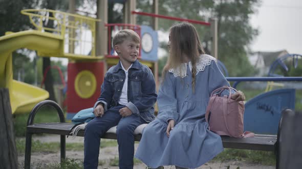 Two Relaxed Positive Children Sitting on Bench on Playground and Talking alt