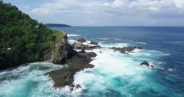 Reveal drone shot of waves of Indian Ocean hitting boulder and hill overgrown by dense of trees besi alt