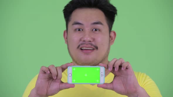 Face of Happy Young Overweight Asian Man Thinking While Showing Phone alt