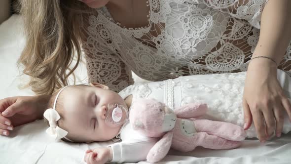 Young Happy Mother Looks at Newborn Sleeping Daughter and Takes Little Hand of Baby Girl