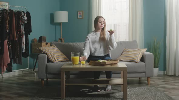 Young Girl in White Sweater Puts Glass of Orange Juice on Table and Bites Pizza