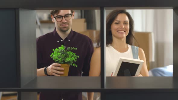 Young Couple in Love Putting Pot with Plant and Photo Frame on Shelves and Hugging alt