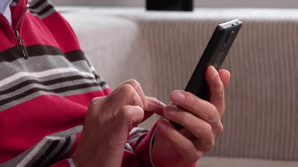 An Elderly Man Sits on a Couch in an Apartment and Works on a Smartphone - Closeup alt