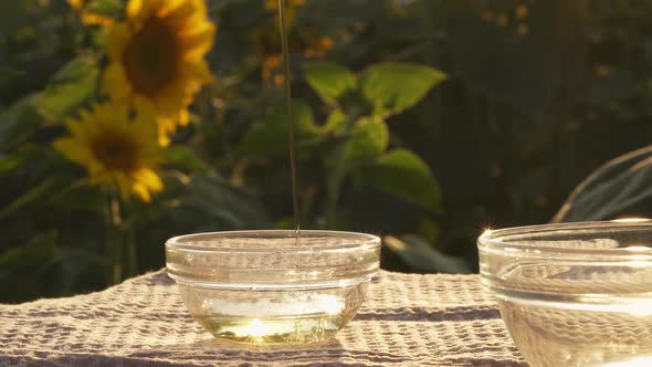 Sunflower Oil Poured In Glass Bowl On Field Of Sunflower Flowers. Oil Is Poured In Bowl In Trickle alt