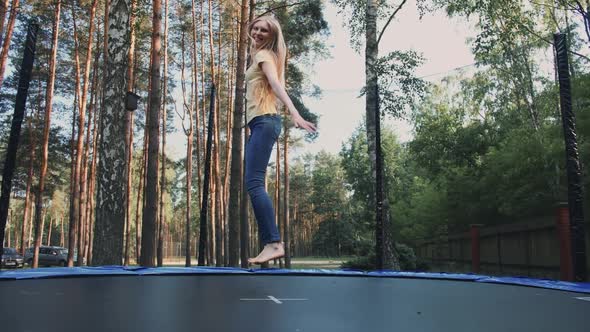 Happy Female Jumping on Trampoline. Beautiful Young Blond Barefoot Woman in Light Summer Shirt and alt