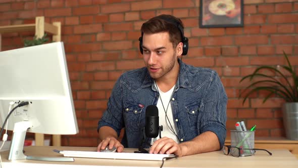 Cheerful Handsome Male Audio Blogger Wearing Headphones with Computer and Professional Microphone alt