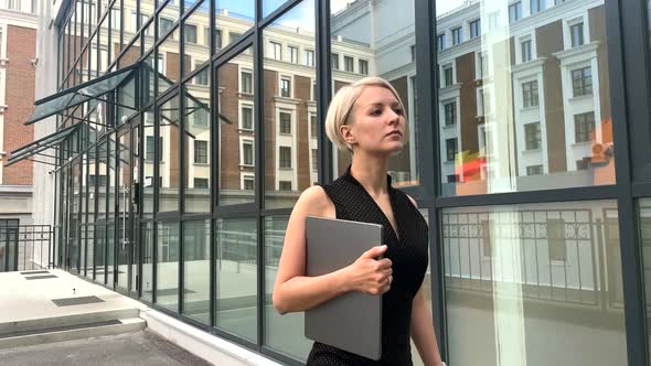 A Young Blonde in a Black Dress with a Tablet Walks Along a Glass Wall alt