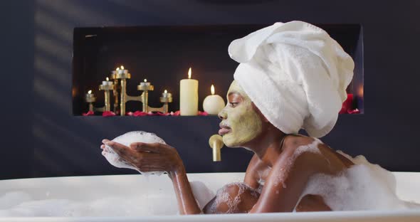 African american woman with towel and mask taking bath and blowing foam in bathroom alt
