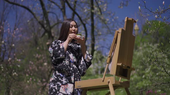 Gorgeous Slim Asian Woman in Kimono Drinking Tea Standing at Easel in Sunny Spring Park Outdoors alt