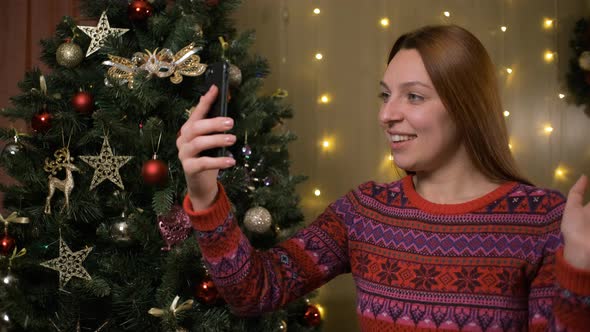 Young Smiling Woman Near Christmas Tree with Making Video Message or Selfie