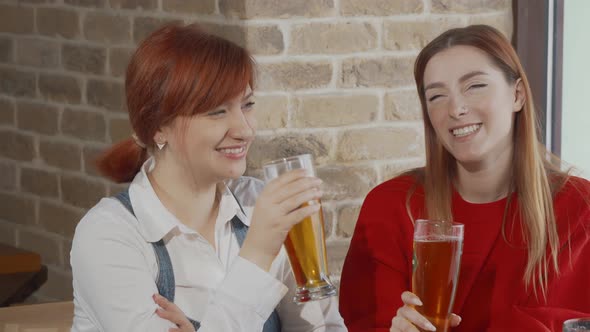 Two Young Women Clinking Beer Glasses Celebrating Together at the Bar
