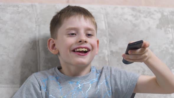 Cheerful caucasian boy 7-8 years old switches channels on holding the tv remote and eats light snack alt