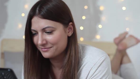 Happy Beautiful Girl in Pajama with Long Dark Hair Enjoys Mobile Phone Lying on Bed at Home and alt