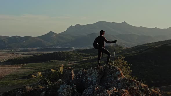 Young Man with a Backpack and Nordic Walking Sticks Stands on the Top of a Mountain Against the