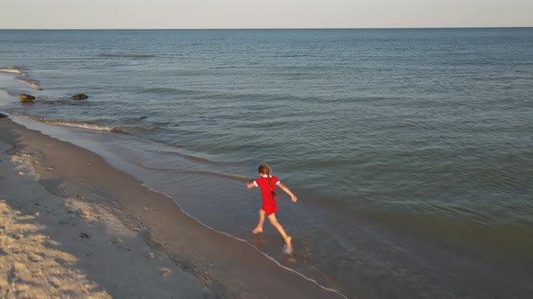 9 Years Old Girl in a Red Dress Runs Along the Water Line on a Deserted Beach
