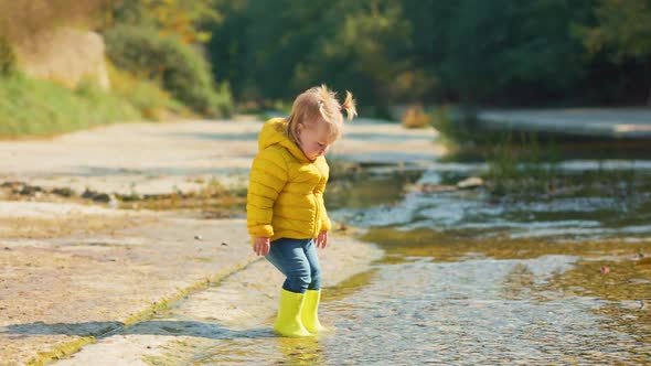 A funny baby girl in yellow rubber boots carefully enters the river and plays with the water alt