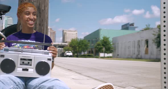 This video is about a young attractive lady sitting against light poll and listening to boom box. Th alt
