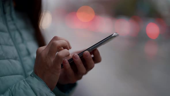 Closeup of a Woman's Hands with a Phone Against the Background of Blurred City Lights alt