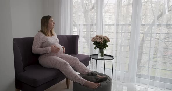 Pregnant Woman Stroking Her Big Belly While Relaxing on the Sofa at Home Near Bouquet of Rose