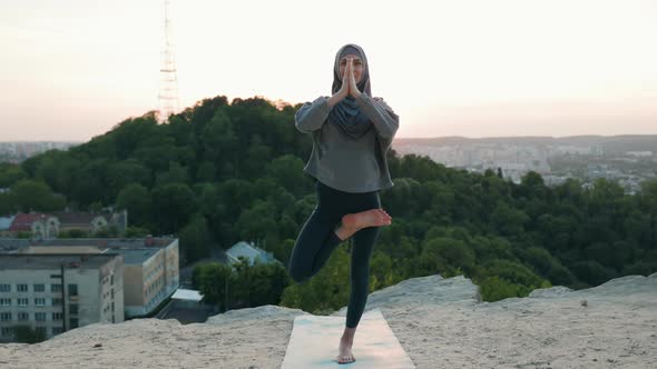 Beautiful Yoga Woman in Hijab on Top of the Hill in the Evening Stays in Tree Position at Sunset