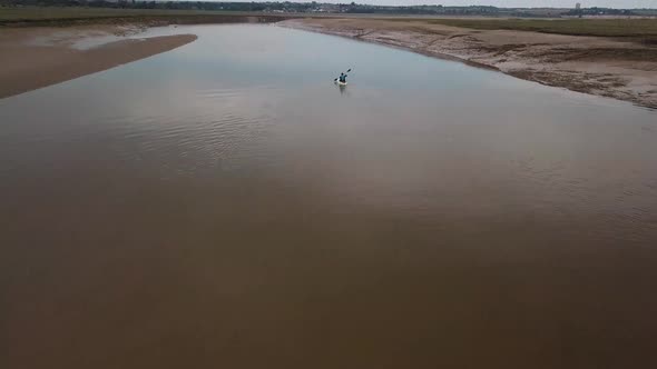 Man kayaking on meandering river, drone pans up to reveal kayaker and beautiful landscape in the bac alt