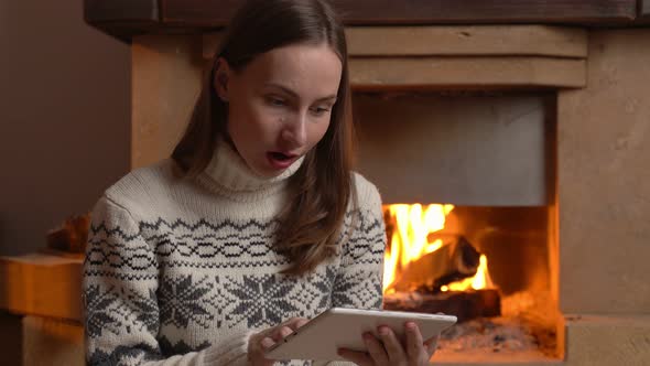 An Excited Woman Holds a Tablet and Celebrates the News While Sitting By the Fireplace alt