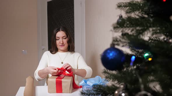 Young Woman Decorates Box with Red Ribbon Next to Christmas Tree alt