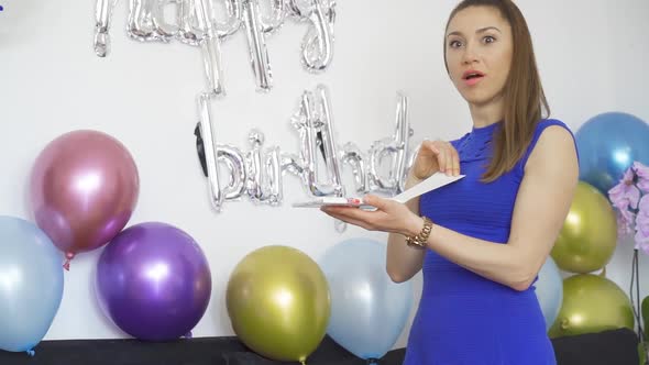 Surprised Young Woman Opening a Birthday Gift at Birthday Party with Colorful Balloons alt
