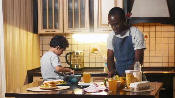 Biracial boy sitting on the table and eating corn flakes near his father