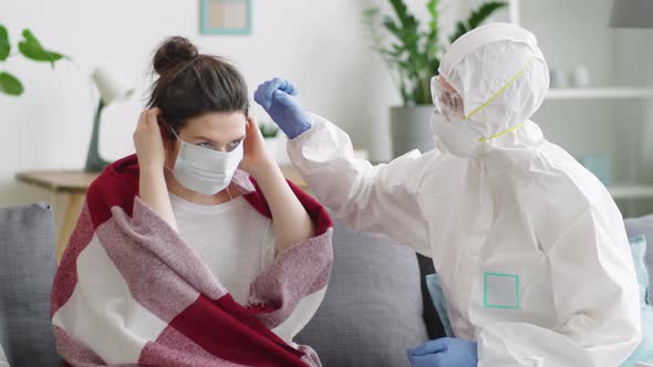 Sick Woman Putting on Face Mask with Help of Doctor in Protective Suit alt