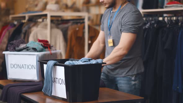 Young Man Volunteer Sorting and Unpacking Clothes in Plastic Boxes alt